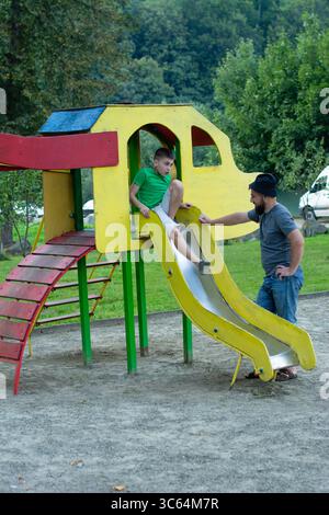 A boy sits at the top of a playground slide as his father assists him. The slide is bright yellow and red. They are in a park-like setting. Stock Photo