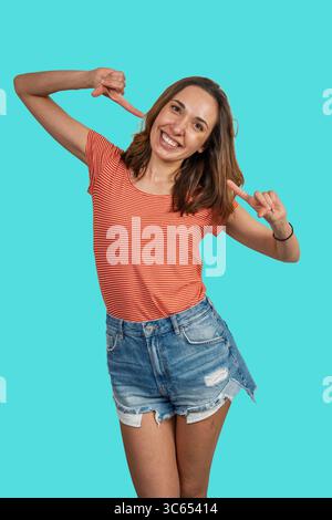 Cheerful young woman pointing herself with both index fingers, smiling and expressing self-confidence and self-promotion, isolated on a vibrant turquo Stock Photo