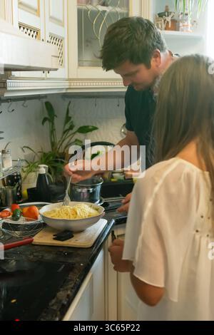 A man and woman are in a bright kitchen, preparing a pasta dish. The man is using a fork to serve the pasta into a bowl, while the woman watches nearb Stock Photo