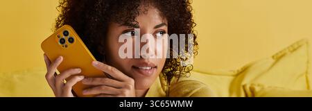 A curly-haired woman enjoys using a smartphone while surrounded by yellow decor, banner Stock Photo