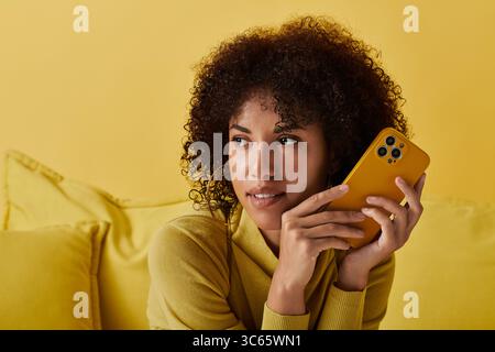A curly-haired woman dressed in a bright yellow outfit holds a smartphone with focus. Stock Photo
