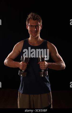 young man with 2x dumbbells in hand, biceps pose, black background Stock Photo