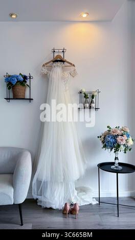 Elegant white wedding dress hanging on a hanger in a softly lit room, prepared for the upcoming marriage celebration Stock Photo