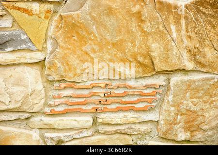 Close-up of a textured stone wall with warm earthy tones and unique red brick inlay, perfect for architectural or background use. Rustic Stone Wall wi Stock Photo