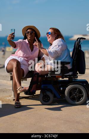 Two smiling women taking a selfie by the beach, with one in a wheelchair Stock Photo