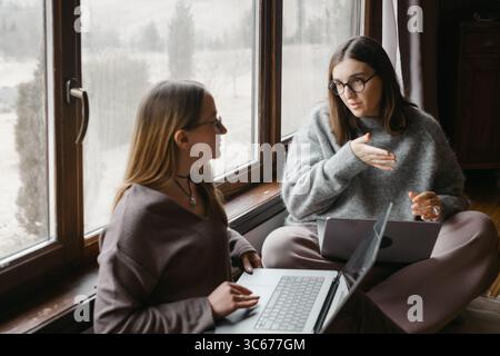 Two women freelancers are sitting near large window, working on their laptop. Friends collaborate or share ideas. Small team communication, work Stock Photo