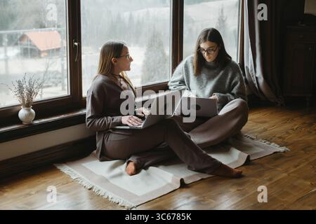 Two women freelancers are sitting near large window, working on their laptop. Friends collaborate or share ideas. Small team communication, work Stock Photo