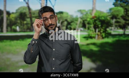 Hispanic man with glasses in a park setting, wearing a dark shirt, showcasing confidence and style beneath the sun-dappled trees on a sunny day. Stock Photo
