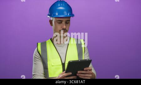 Young man in reflective vest and hardhat examines a tablet against isolated purple background, symbolizing technology and construction with a professi Stock Photo