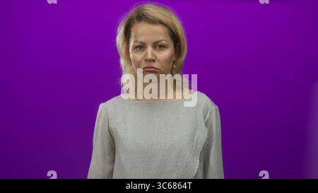 Young blonde woman pouting lips in purple studio setting wearing gray blouse, facing camera with a sullen expression; disappointment. Stock Photo