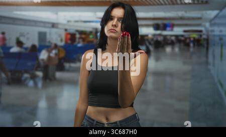 Young hispanic woman beckons with open hand at crowded airport terminal; delayed flight impatience. Stock Photo