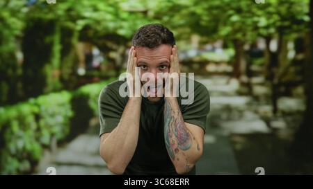 Young man with tattoos and short hair expressing stress in a lush green park under dappled sunlight. Stock Photo