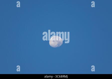 A beautiful, nearly full moon shines brightly in a clear blue sky during the day. Stock Photo