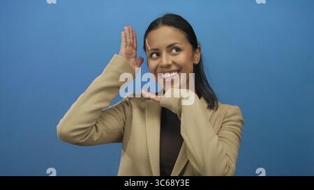 Woman smiling against blue background with hands framing face wearing beige jacket representing happiness and youthful energy in an isolated studio se Stock Photo