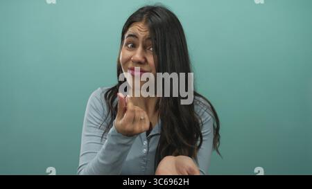 Young hispanic woman beckons open palm for money in studio against green wall, making beckoning gesture; urgent plea. Stock Photo