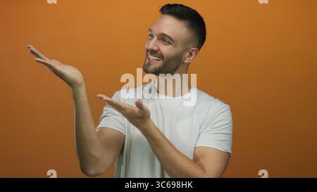 Young hispanic man smiling and extending arms to present something against a bright orange background, showcasing a welcoming gesture with joyful expr Stock Photo