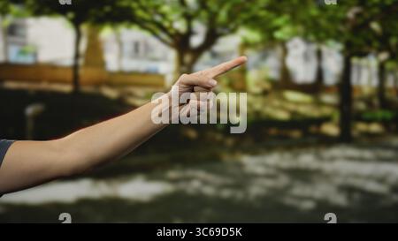Man's hand pointing forward in a lush green park setting, showcasing natural daylight and a calm outdoor atmosphere with trees and shadows surrounding Stock Photo