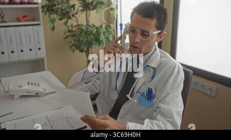 Young hispanic male doctor with stethoscope in hospital room talking on phone while reviewing documents at his desk. Stock Photo