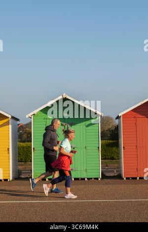 England, Sussex, East Sussex, Eastbourne, Colourful Beach Huts on the Beachfront, Elderly Couple Jogging Stock Photo