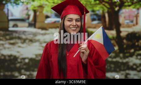 Woman in red graduation gown holding philippine flag in a park setting, symbolizing accomplishment, pride, and national identity. Stock Photo