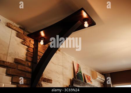 Modern wooden ceiling fixture with bulbs, mounted on a stone wall inside a stylish interior. Interior of the apartment, the details of the lighting. Stock Photo