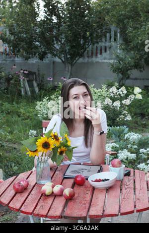 Smiling woman artist with tablet eating berries while sitting at a red round table in garden with tablet and apples on white background. Idea of onlin Stock Photo