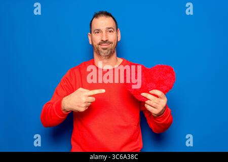 Bearded Hispanic man in his 40s wearing a red sweater holding a red heart as a symbol of romance and health, looking surprised and pointing at himself Stock Photo