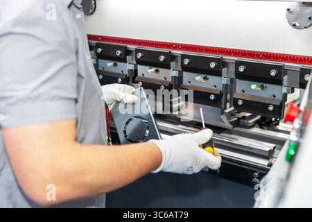 A close-up view of a factory worker operating a CNC press brake machine while bending a sheet metal component. The technician is wearing white safety Stock Photo
