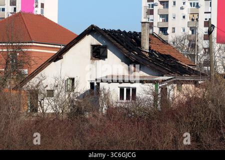 A white house with extensive fire damage and a partially collapsed roof sits abandoned in an urban setting, contrasted by nearby modern buildings Stock Photo