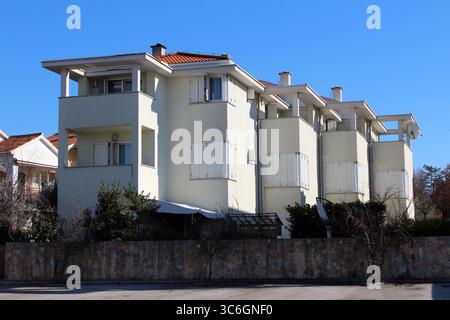 A large, modern apartment building with clean white walls, red tiled roof, and closed white shutters sits quietly behind a low stone wall in a green Stock Photo