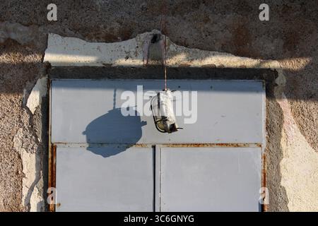 A worn industrial lamp with metal cage hangs by a cord, casting a distinct shadow on a sunlit, cracked concrete wall above a rust-stained metal door Stock Photo