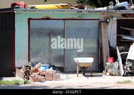 A metal-clad garage stands among scattered bricks, wheelbarrow, wood, and tools, surrounded by various construction materials in an active, sunlit Stock Photo