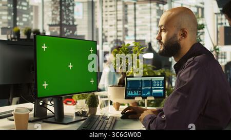 Man in office reviewing financial graphs on green screen PC, analyzing key performance indicators. Employee looking over data trends and strategic insights on chroma key computer, camera B Stock Photo