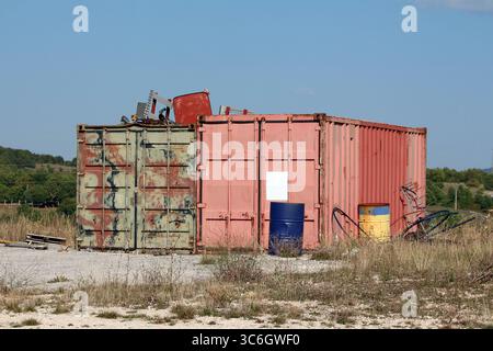 Two old shipping containers, one faded red and one distressed green, stand on gravel with blue and yellow barrels, metal scrap, and overgrown grass Stock Photo