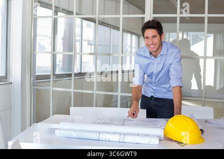 Male architect in twenties reviewing blueprints at office desk with yellow hard hat, copy space. Professional, urban, modern, creative, collaborative, Stock Photo