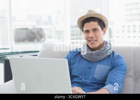 Man wearing straw hat, gray scarf sitting on sofa by windows using laptop, copy space. Casual, modern, bright, urban, lifestyle, relaxation, technolog Stock Photo
