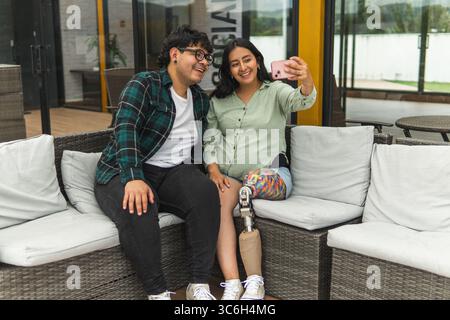 Smiling man and woman with prosthetic leg taking selfie with smartphone while relaxing on outdoor sofa Stock Photo
