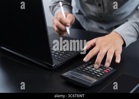 Close up of man's hands using a calculator and laptop while holding a digital pen. Ideal for finance, budgeting, tax planning, accounting, audit, or d Stock Photo