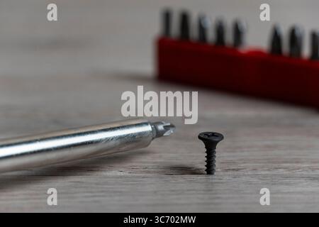 Close-up of a screwdriver and screw resting on a wooden surface, with a blurred red holder of interchangeable bits in the background Stock Photo