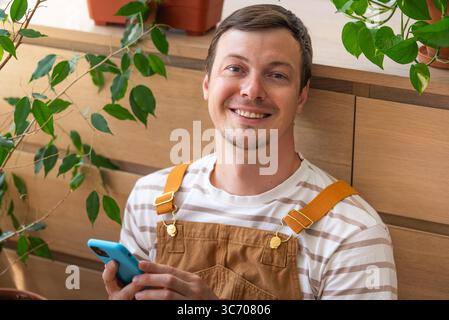 Cheerful gardener surrounded by verdant houseplants, scrolling smartphone screen with satisfied expression. Comfortable home workspace merging professional tasks with botanical enthusiasm Stock Photo