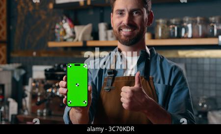 Satisfied happy Caucasian barista man showing green screen mobile phone thumb up in cafeteria cafe male small business entrepreneur recommend Stock Photo