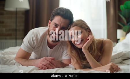Carefree family glad Caucasian couple relaxing in bed looking at camera in bedroom pair wife husband lying cozy morning tenderness married woman man Stock Photo
