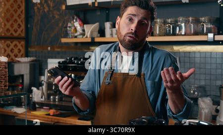 Confused Caucasian barista man holding mobile phone shrug shoulders in cafeteria guy waiter unsure worker looking screen puzzled male bartender Stock Photo