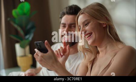 Happy Caucasian couple family using mobile phone together in bedroom boyfriend girlfriend laughing pair scroll smartphone woman man purchasing goods Stock Photo