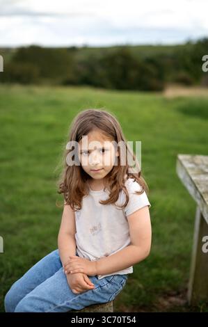 Child girl sitting on a wooden bench in a countryside field. Stock Photo