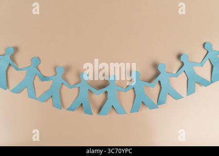 Blue paper cutout of people holding hands in a line on beige background. Symbol of unity, teamwork, population, social connection, and community Stock Photo