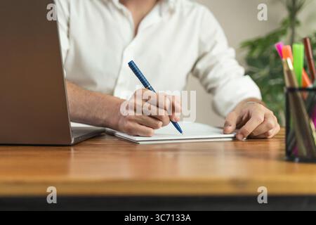 An anonymous professional man in a white shirt sits at a wooden desk with a laptop and writes in a notebook, signing a document or taking notes during Stock Photo