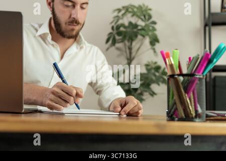 A focused man sits at his desk with a laptop and writes down ideas or signs a document in a notebook, with a shallow depth of field focusing on his ha Stock Photo