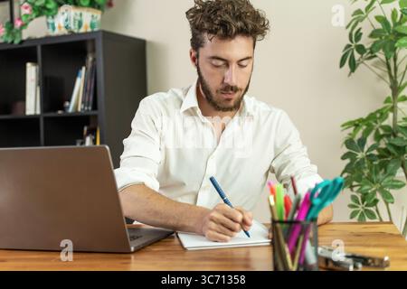 A handsome young student or freelancer with a beard concentrates as he writes in a journal, studying for an exam or planning a project in his home off Stock Photo