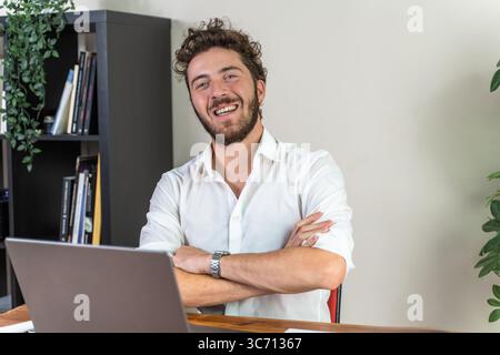 Portrait of a happy and confident young businessman with a beard, sitting with arms crossed at his desk with a laptop and smiling directly at the came Stock Photo